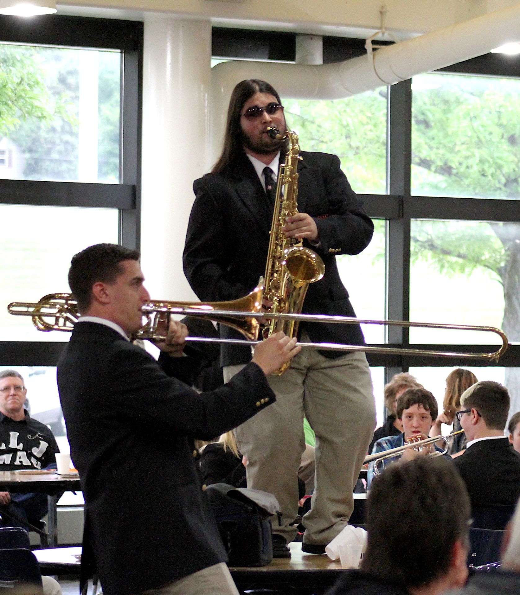 Noah Brant standing on table playing saxophone with trombonist and audience members
